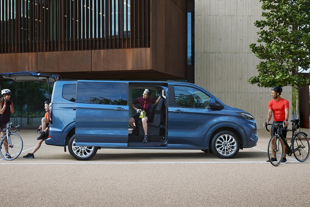 Een blauwe Ford Tourneo Custom, geparkeerd met zijn zijdeuren open en mensen met fietsen in de buurt.