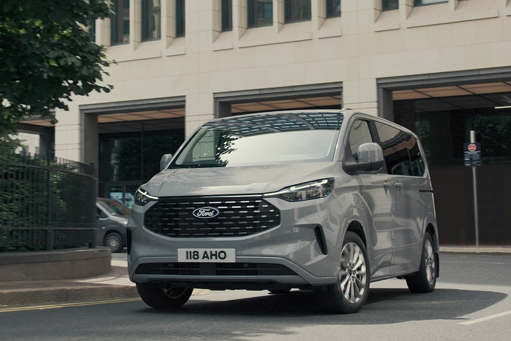 Een zilverkleurige Ford Tourneo Custom rijdt op een stadsweg.