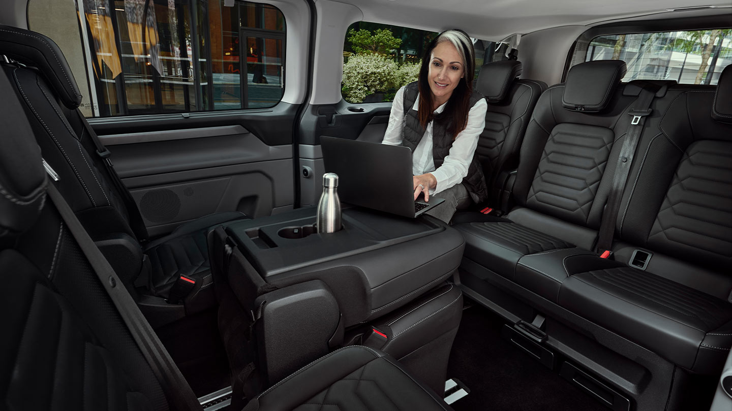 Een vrouw zit in een Ford Tourneo Custom aan een tafel te werken op een laptop.