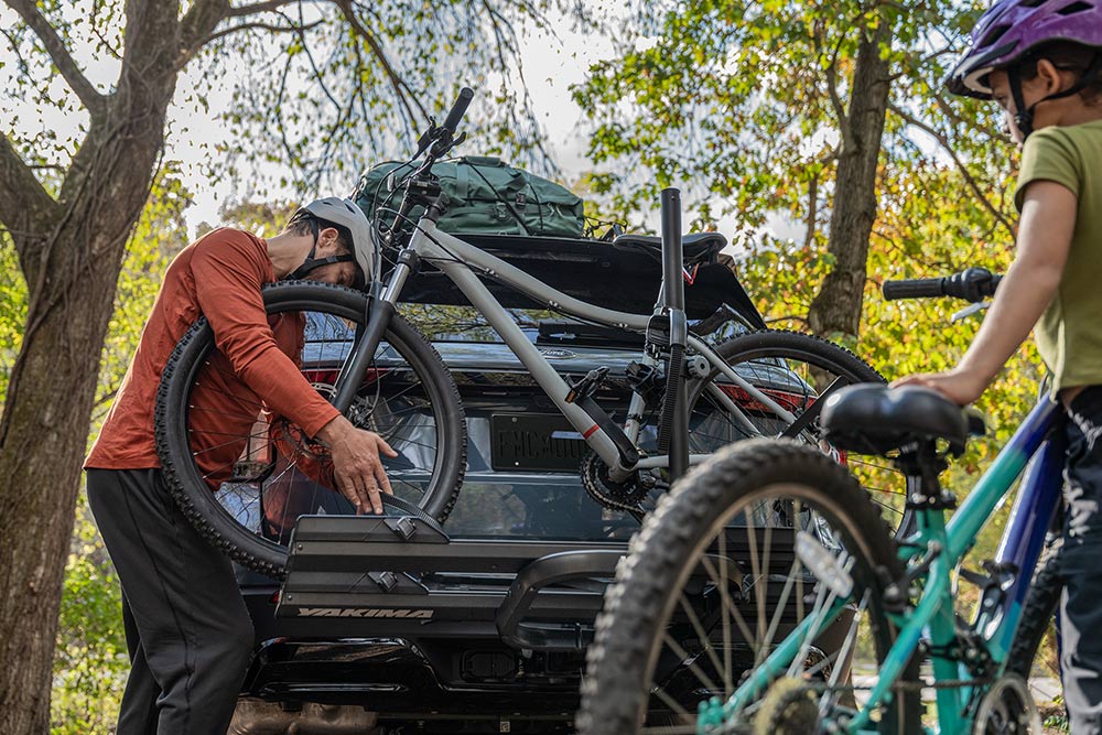 Een persoon laadt een fiets op de achterste fietsendrager van een auto, met een kind en een andere fiets in de buurt. 