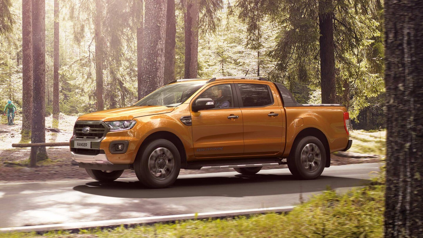 De zon schijnt op de Oranje Ford Ranger Wildtrak die op een bosweg geparkeerd staat, zijaanzicht