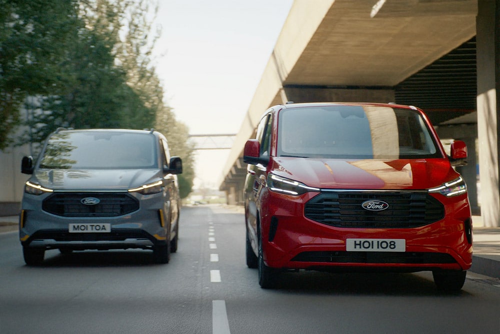 Grijze en rode Ford Transit Custom bestelwagens rijden langs een snelweg.