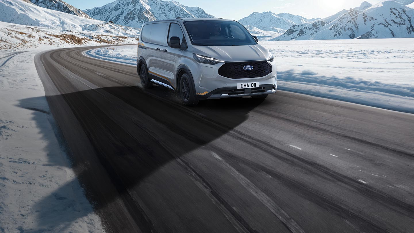 Een zilverkleurige Ford E-Transit Custom die over een besneeuwde bergweg rijdt onder een helderblauwe lucht.