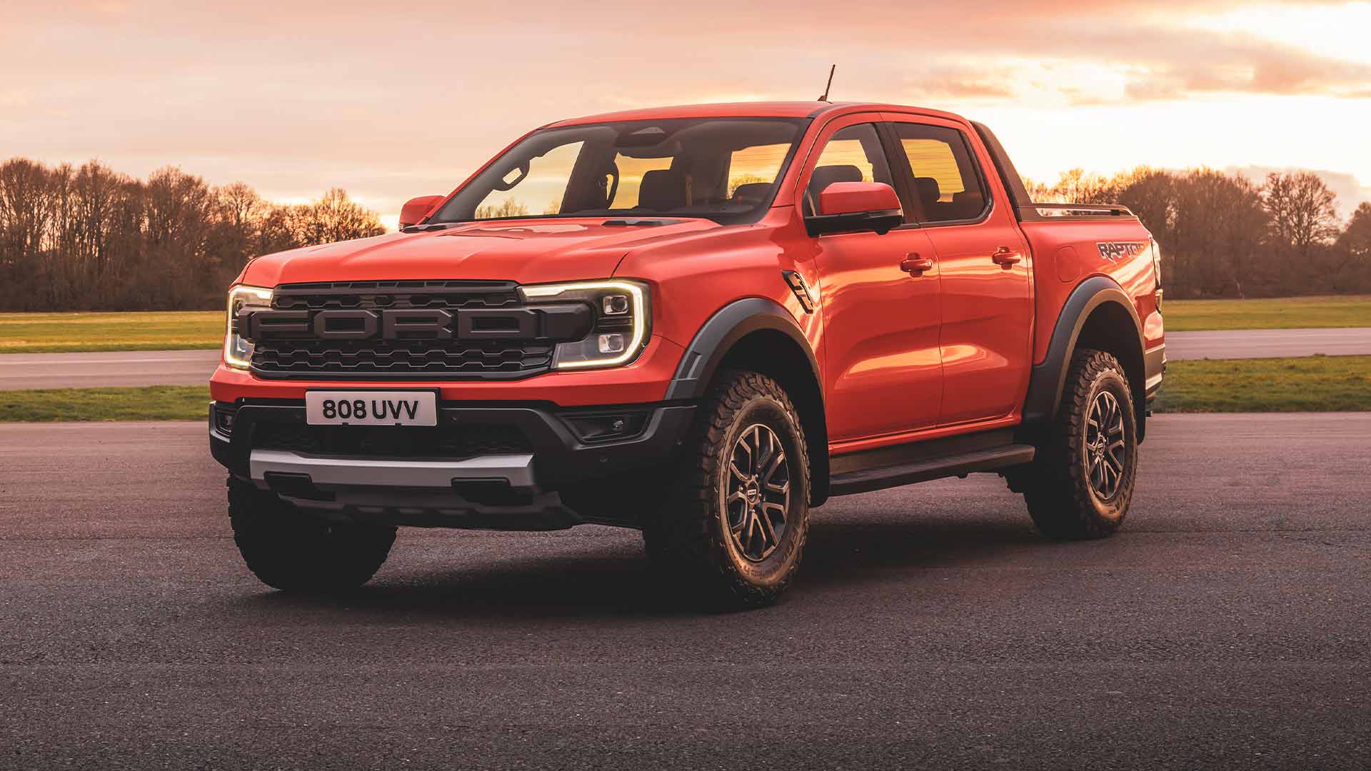 Ford Ranger Raptor in de kleur Code Orange geparkeerd op een circuit met koplampen aan. 