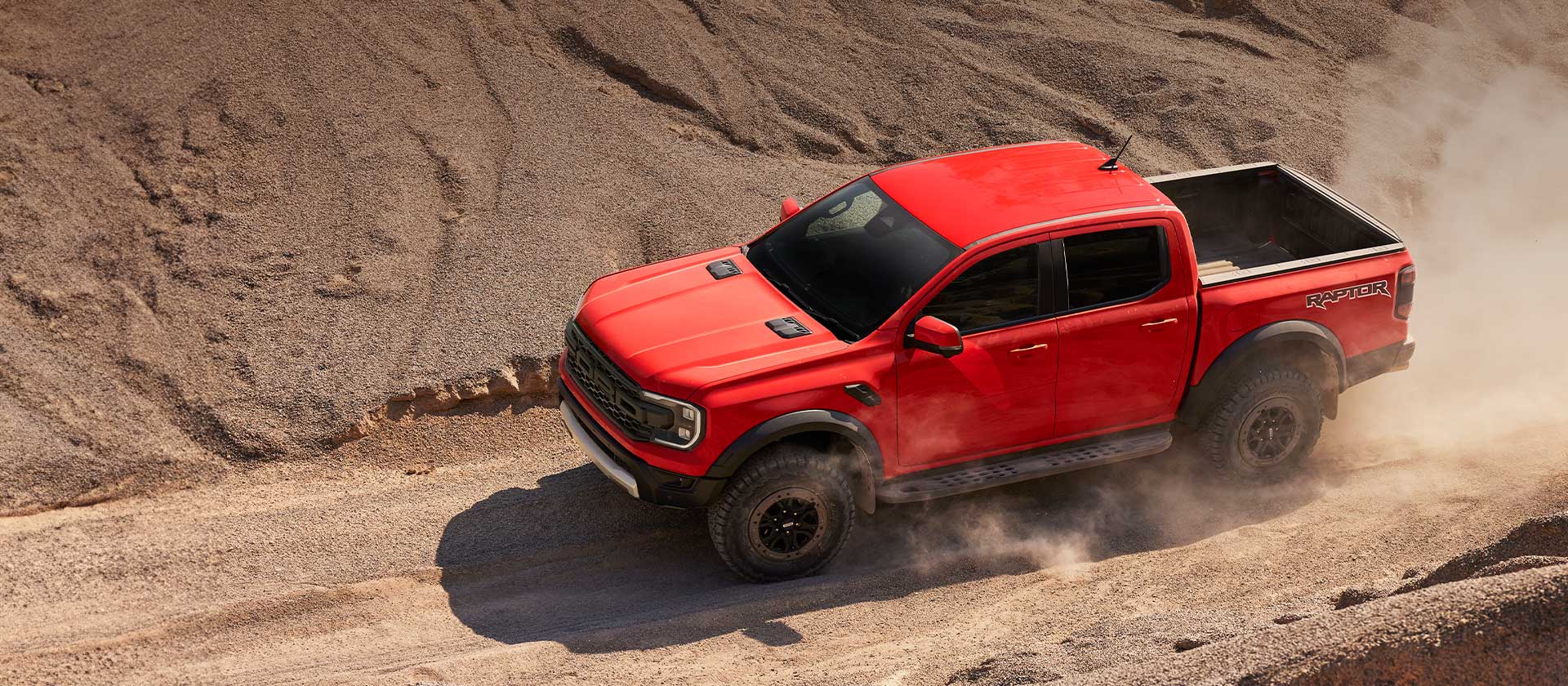 Oranje Ford Ranger Raptor rijdt door stof op een heuvel.