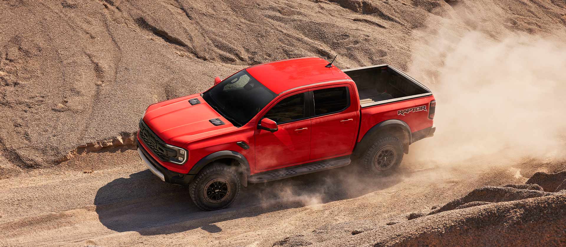 Oranje Ford Ranger Raptor rijdt door stof op een heuvel.