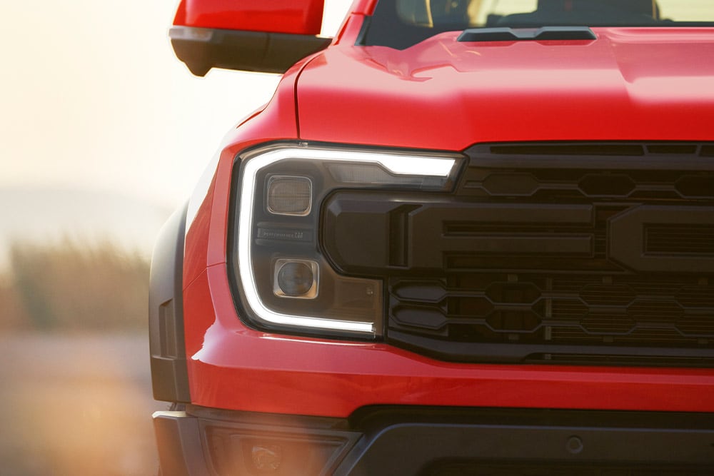 Close-up van een koplamp en grille van een Ford Ranger Raptor in de kleur Code Orange.