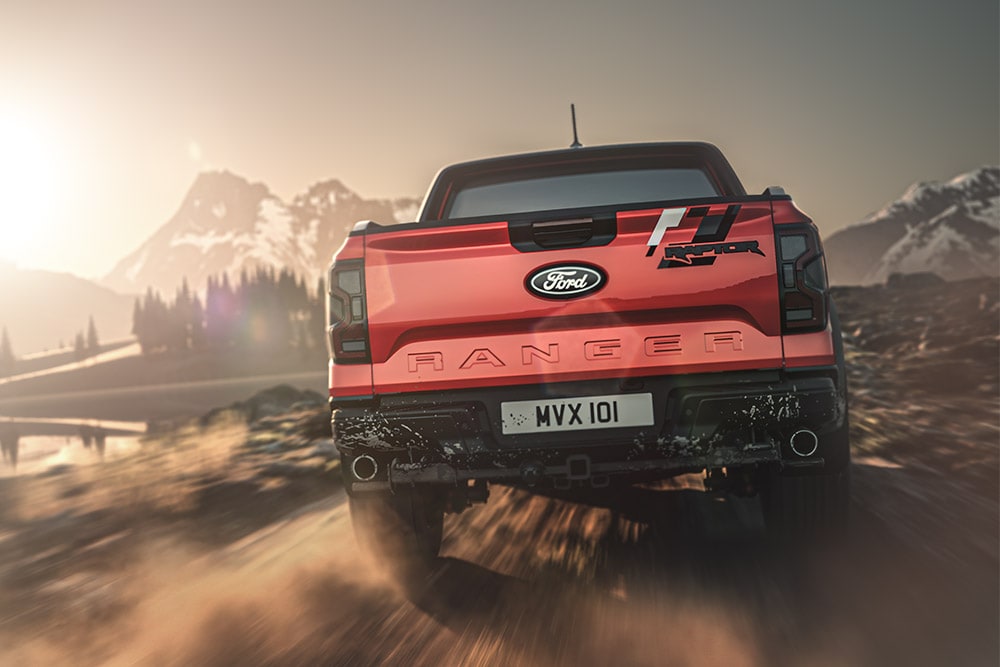 Achteraanzicht van een rode Ford Ranger Raptor die over een onverharde weg rijdt, met bergen op de achtergrond.