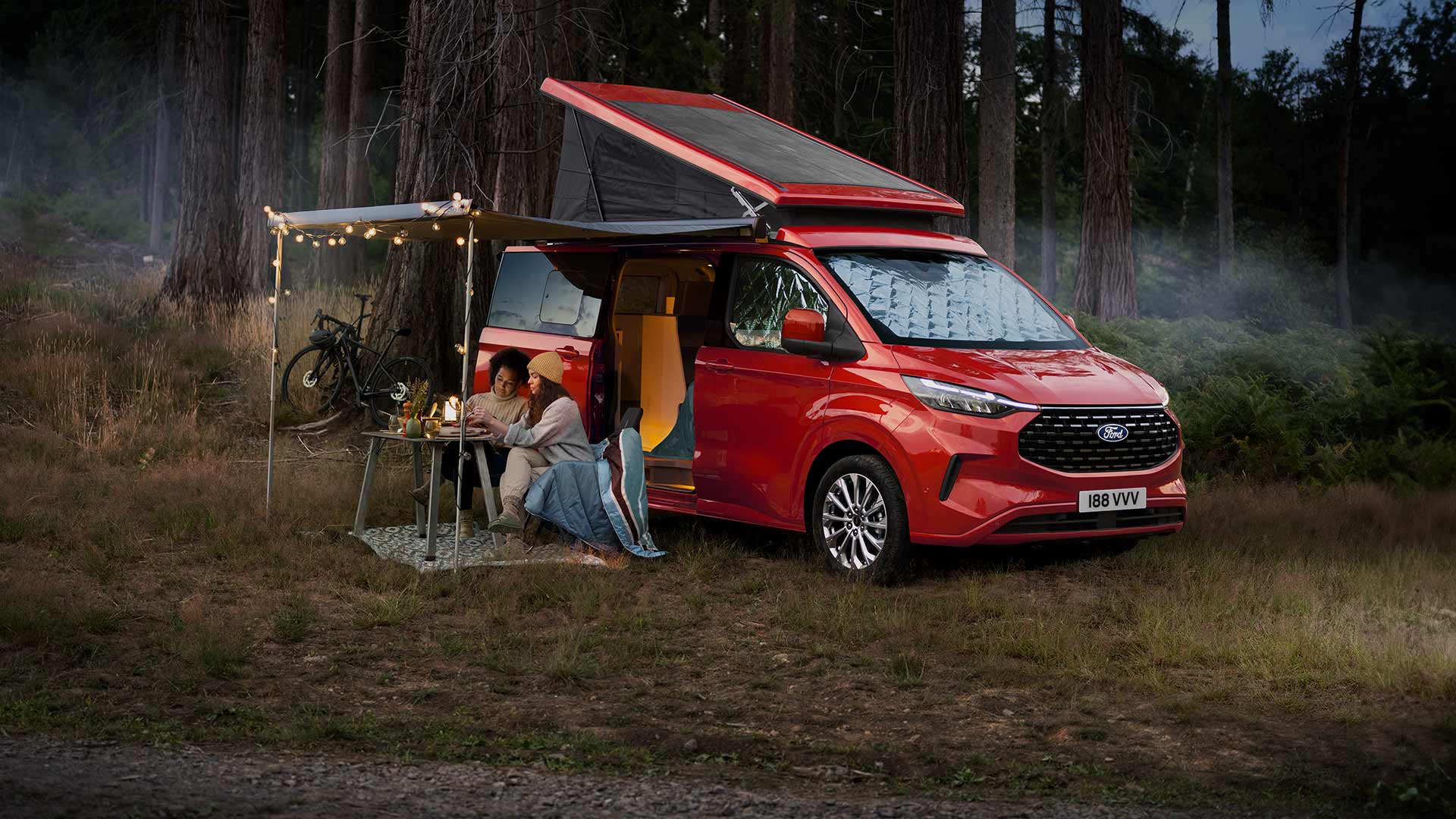 Twee vrouwen zitten onder een luifel van de open zijdeuren van een rode Ford Transit Custom Nugget, met het kanteldak omhoog.