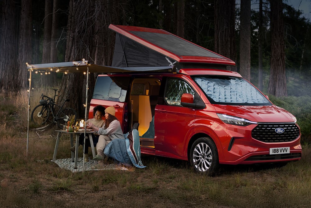 Twee vrouwen zitten onder een luifel van de open zijdeuren van een rode Ford Transit Custom Nugget, met het kanteldak omhoog.