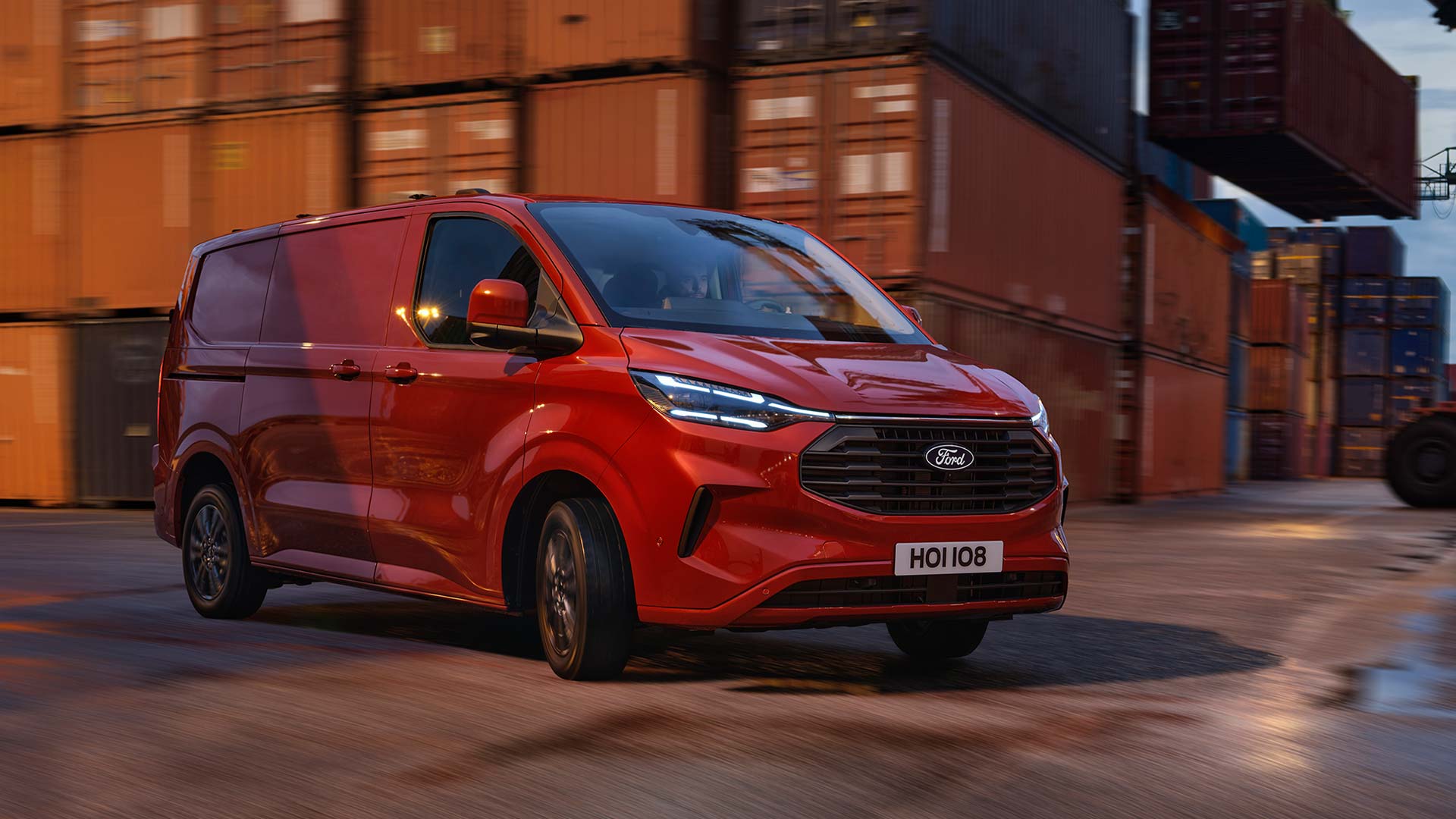 Een rode Ford Transit Custom bestelwagen rijdt door een scheepswerf. 