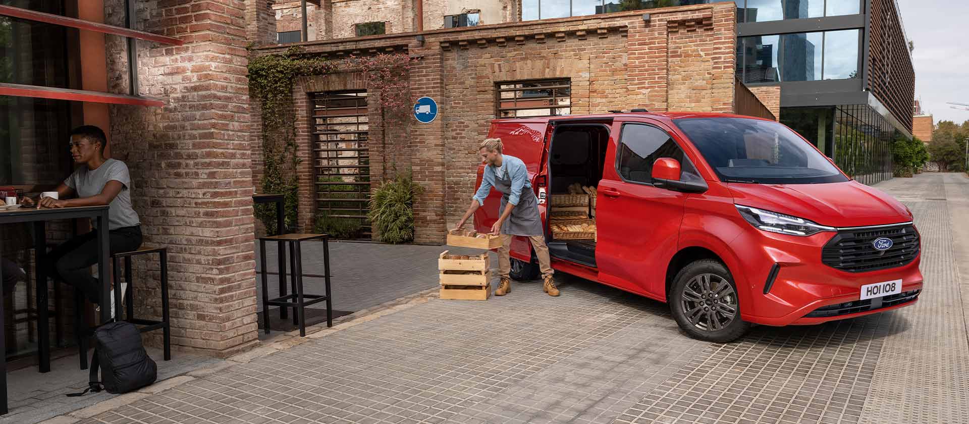 Een persoon laadt kratten uit een rode Ford Transit Custom bestelwagen die voor een coffeeshop staat geparkeerd. 