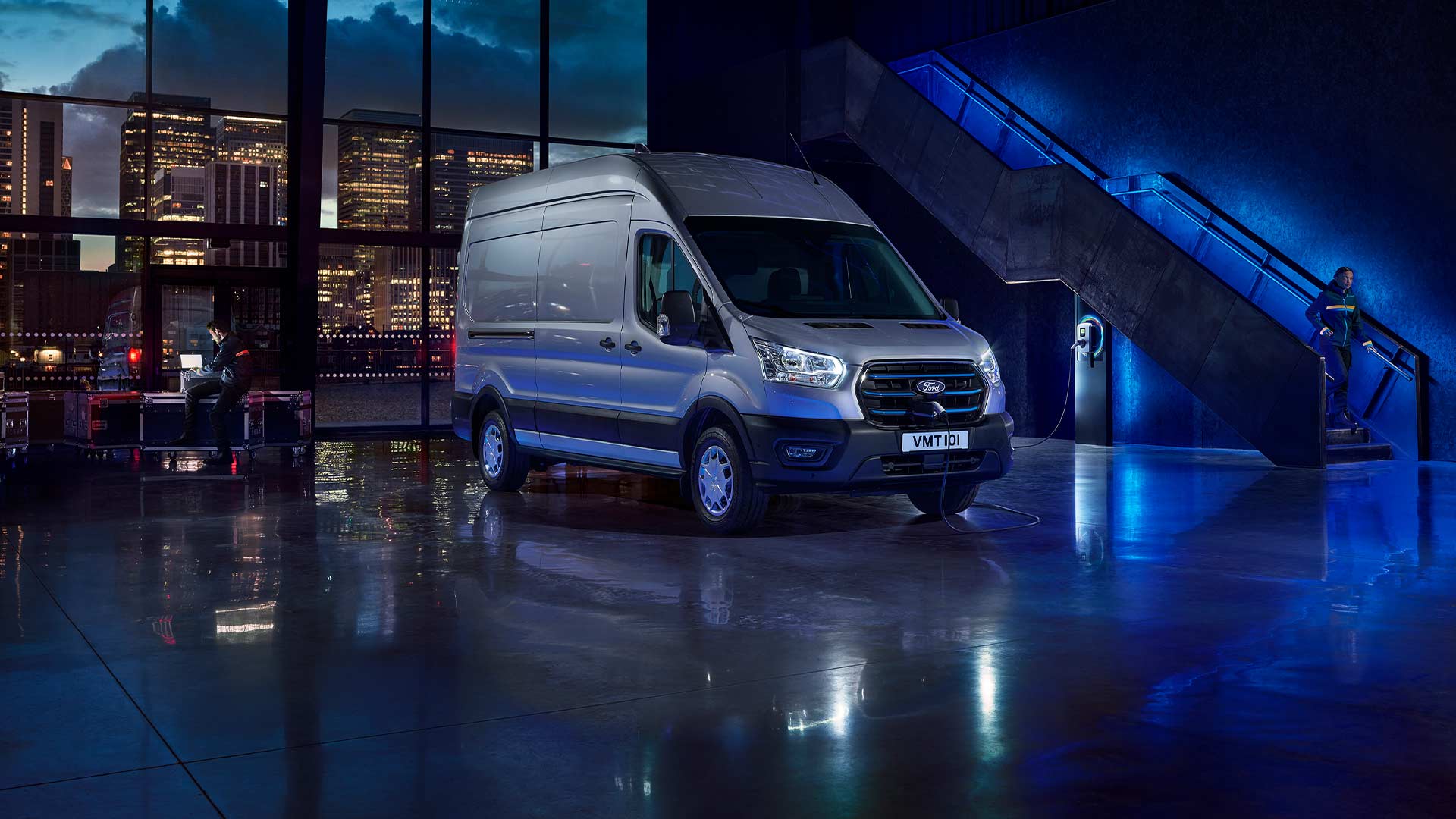 Zilverkleurige Ford E-Transit elektrische bestelwagen overdekt geparkeerd met een skyline van de stad door grote ramen.