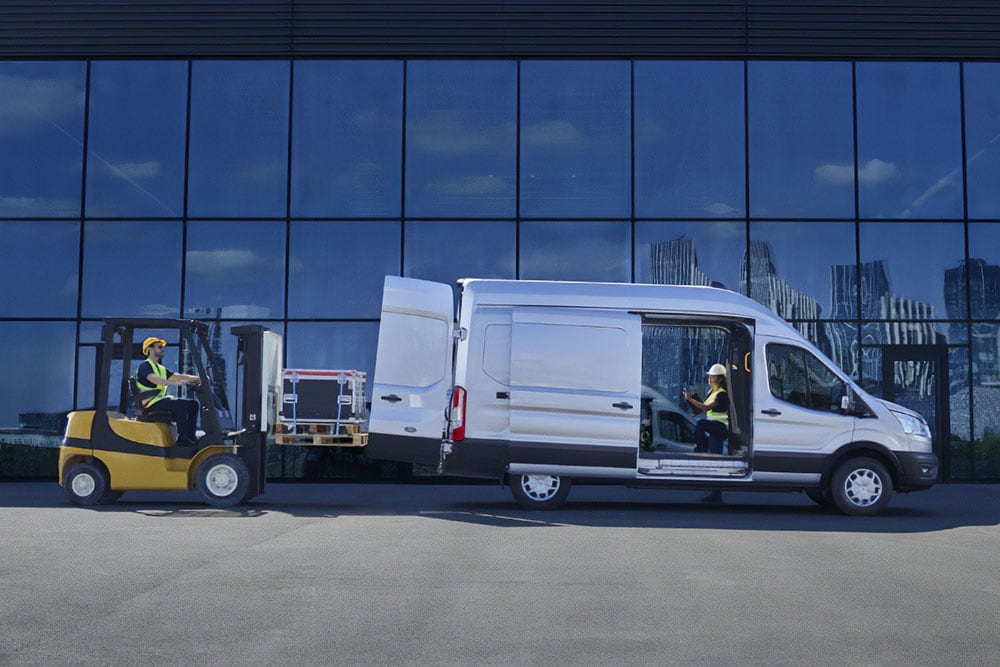 Twee mensen laden vracht in een zilverkleurige Ford Transit bestelwagen met een vorkheftruck.
