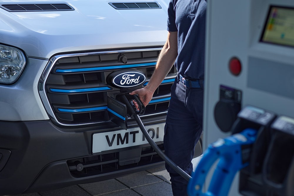 Close-up van een persoon die een oplaadkabel aansluit op een zilverkleurige Ford E-Transit elektrische bestelwagen.