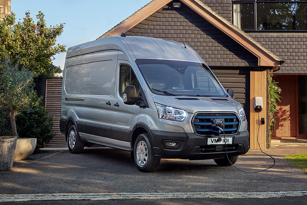 Zilverkleurige Ford E-Transit bestelwagen geparkeerd op een oprit naast een huis, aangesloten op een laadpaal.