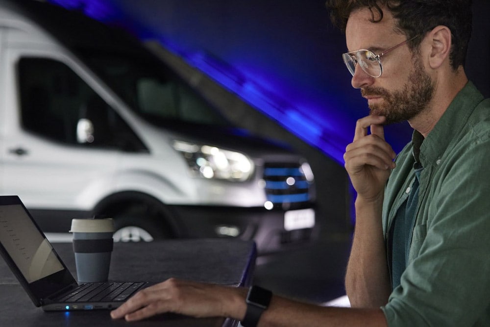 Man werkt op een laptop met een Ford E-Transit elektrische bestelwagen op de achtergrond.