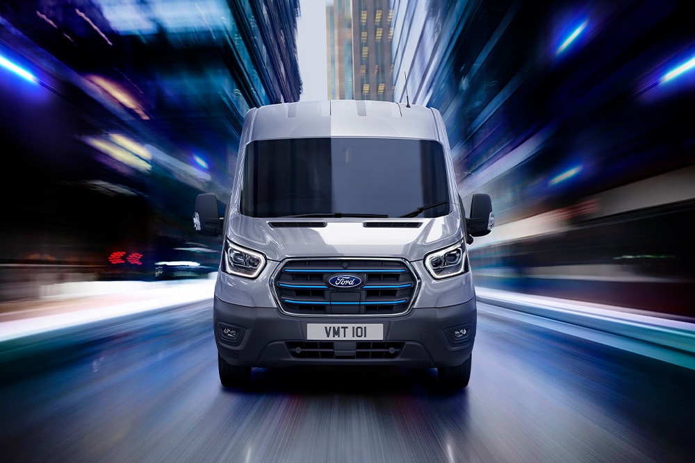 A vooraanzicht van een grijze Ford Transit die 's nachts met snelheid door een stad rijdt, met blauwe grille-accenten