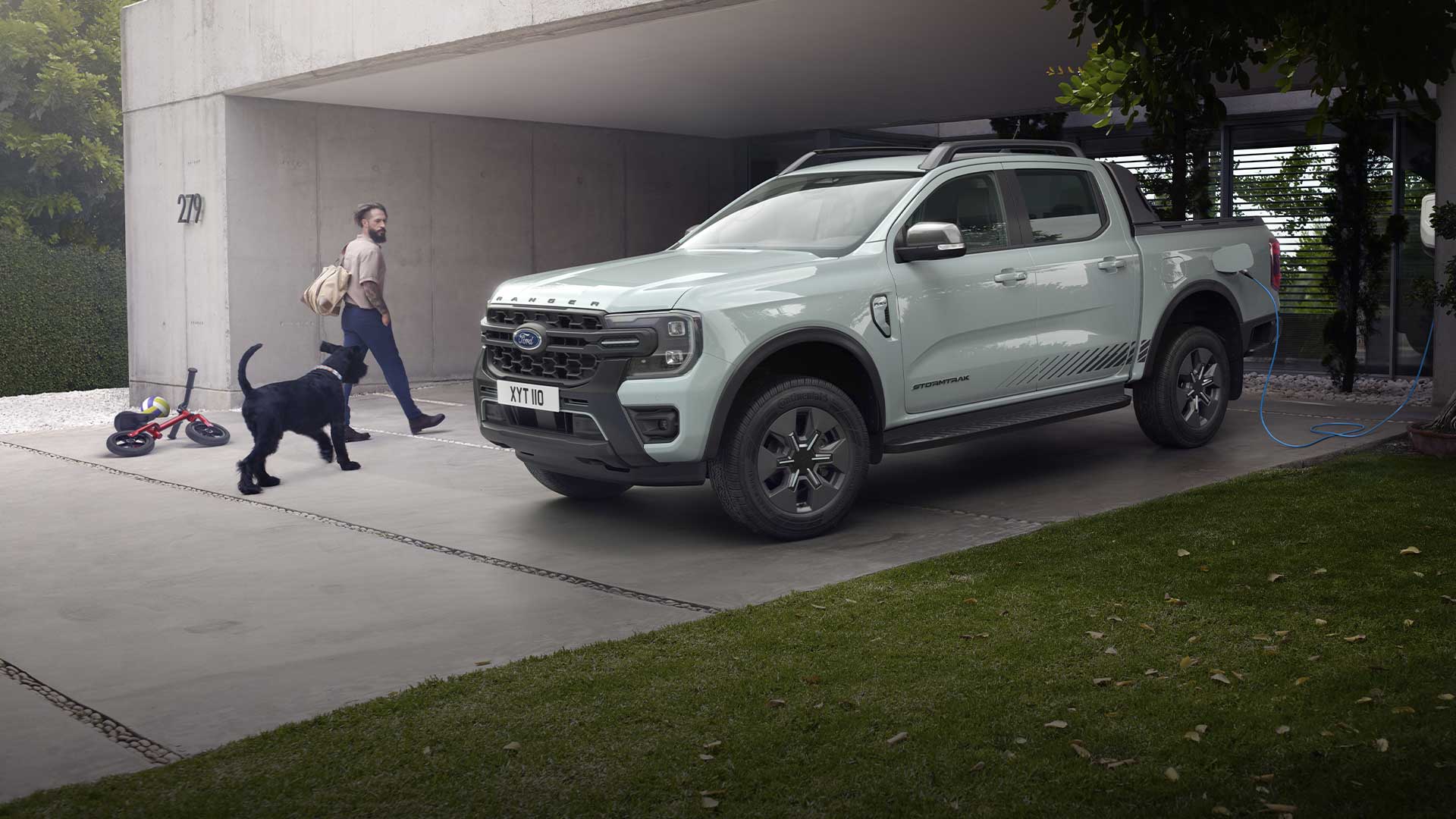 Een man en een hond lopen naar een grijze Ford Ranger plug-in hybride pick-up die op een oprit staat te laden. 