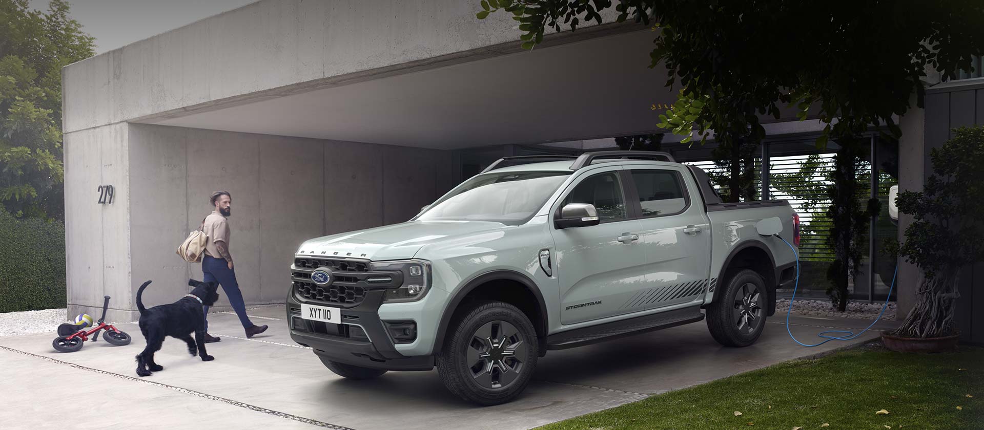 Een man en zijn hond gaan een modern huis binnen en wandelen langs een Ford Ranger Stormtrak PHEV tijdens het laden.