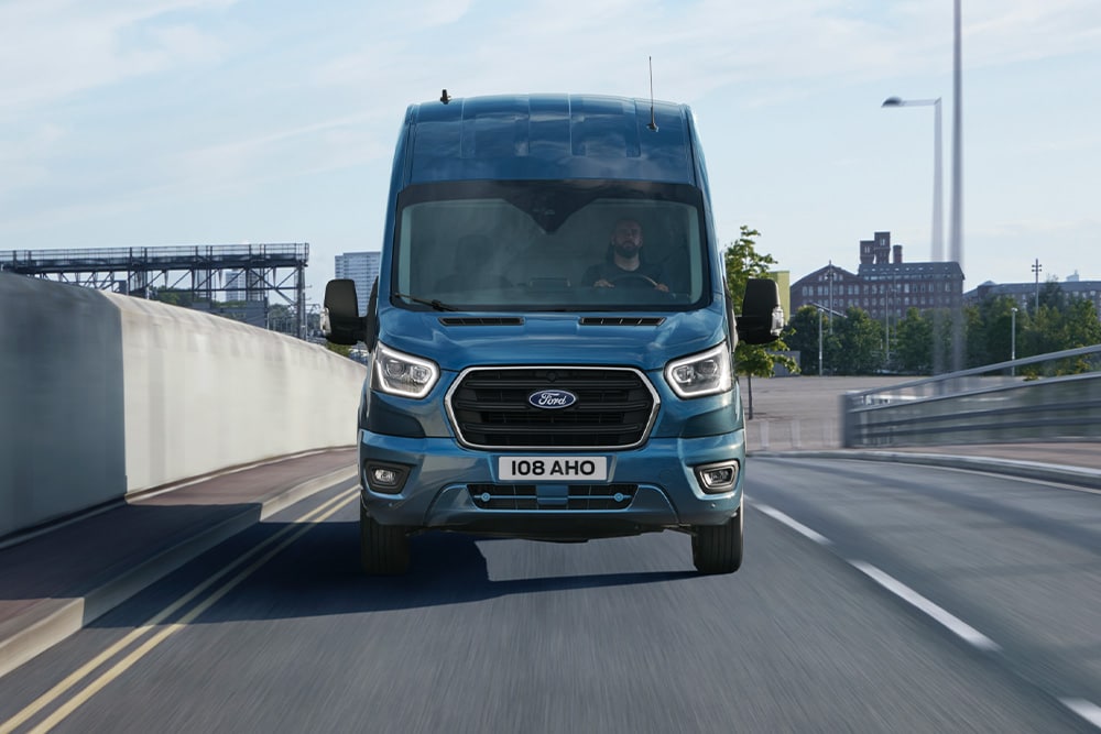 Vooraanzicht van blauwe Ford Transit bestelwagen rijdend op een weg. 