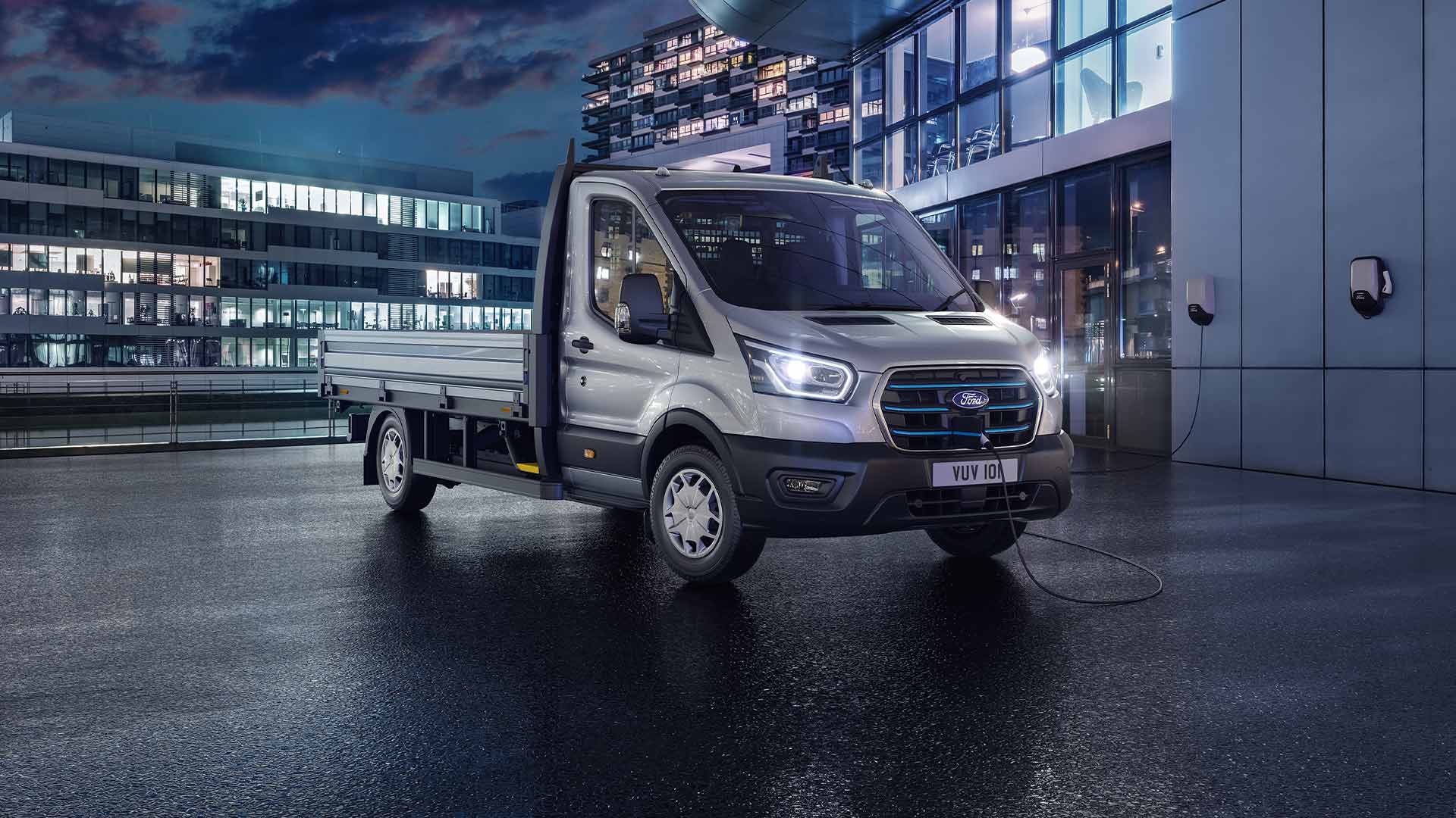 Zilverkleurige Ford Transit Chassis Cabine geparkeerd voor een modern gebouw in het donker.