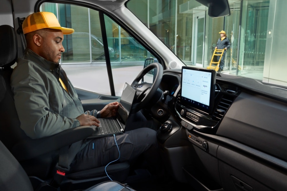 Een man werkt op een laptop in de bestuurdersstoel van een Ford Transit bestelwagen.