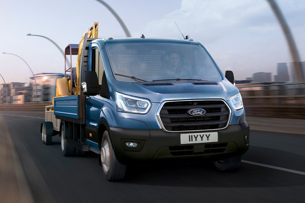 Vooraanzicht van de grille van een Ford Transit Chassis Cab die met een graafmachine over een stadsweg rijdt. 