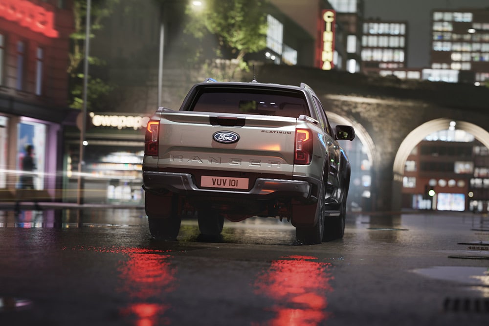 Achteraanzicht van een zilverkleurige Ford Ranger die in het donker door een neonverlicht stadslandschap rijdt.