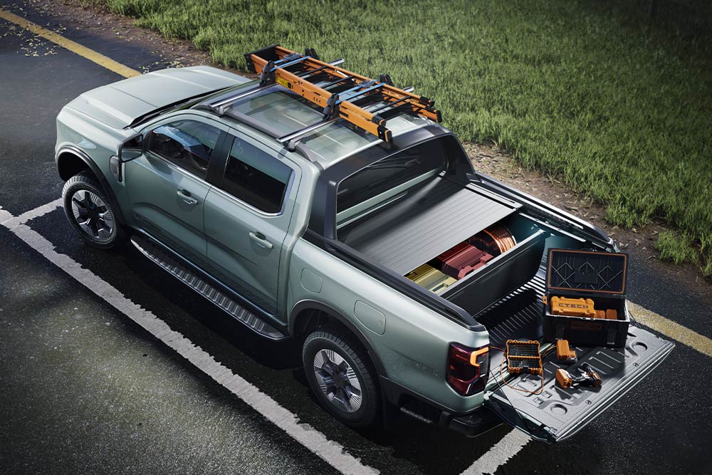 Achteraanzicht vanuit een hoge hoek van een grijze Ford Ranger pick-up truck, met open laadbak vol gereedschap en een dakdrager op een verharde weg.