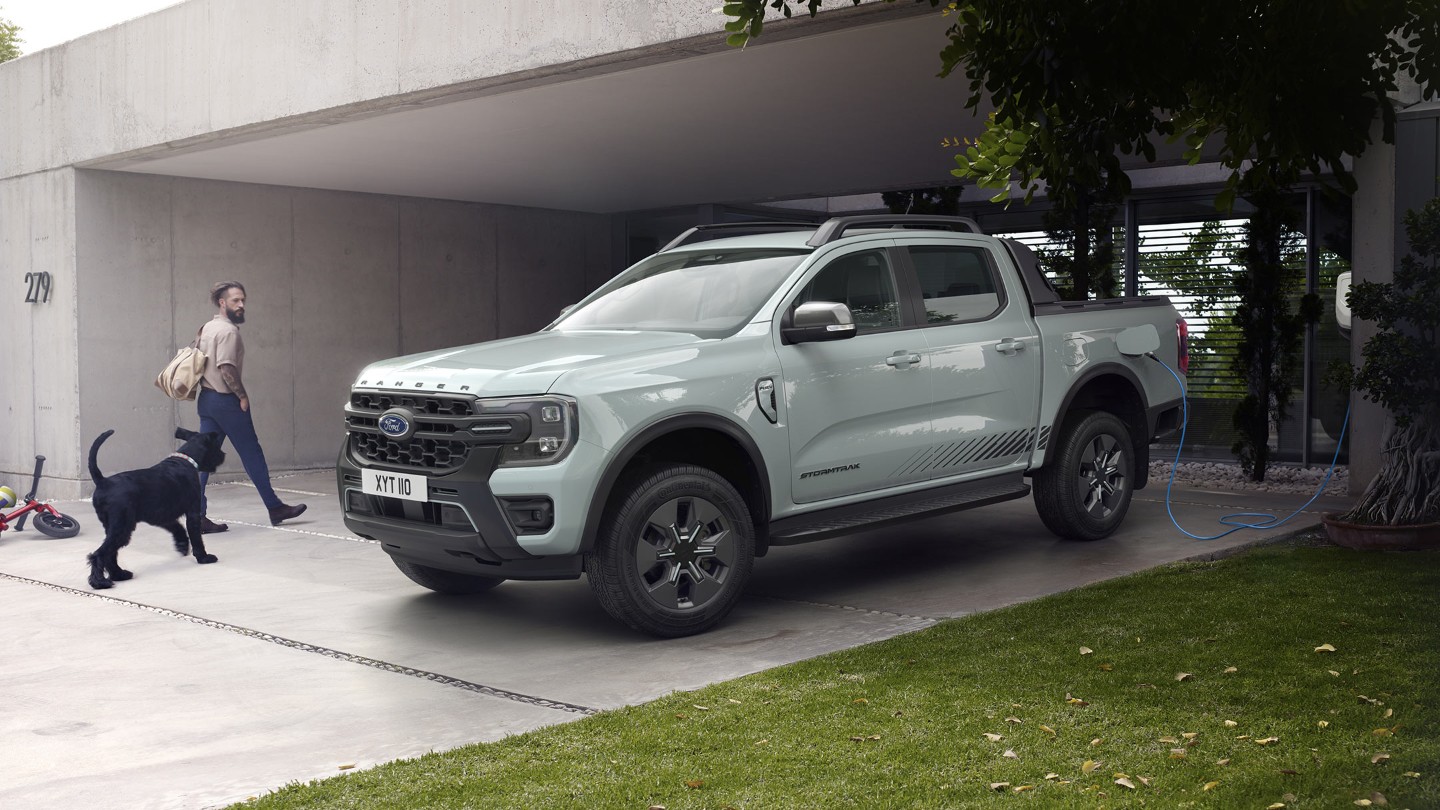 Een man en zijn hond gaan een modern huis binnen en wandelen langs een geparkeerde Ford Ranger Stormtrak Plug-in Hybrid die wordt opgeladen. 