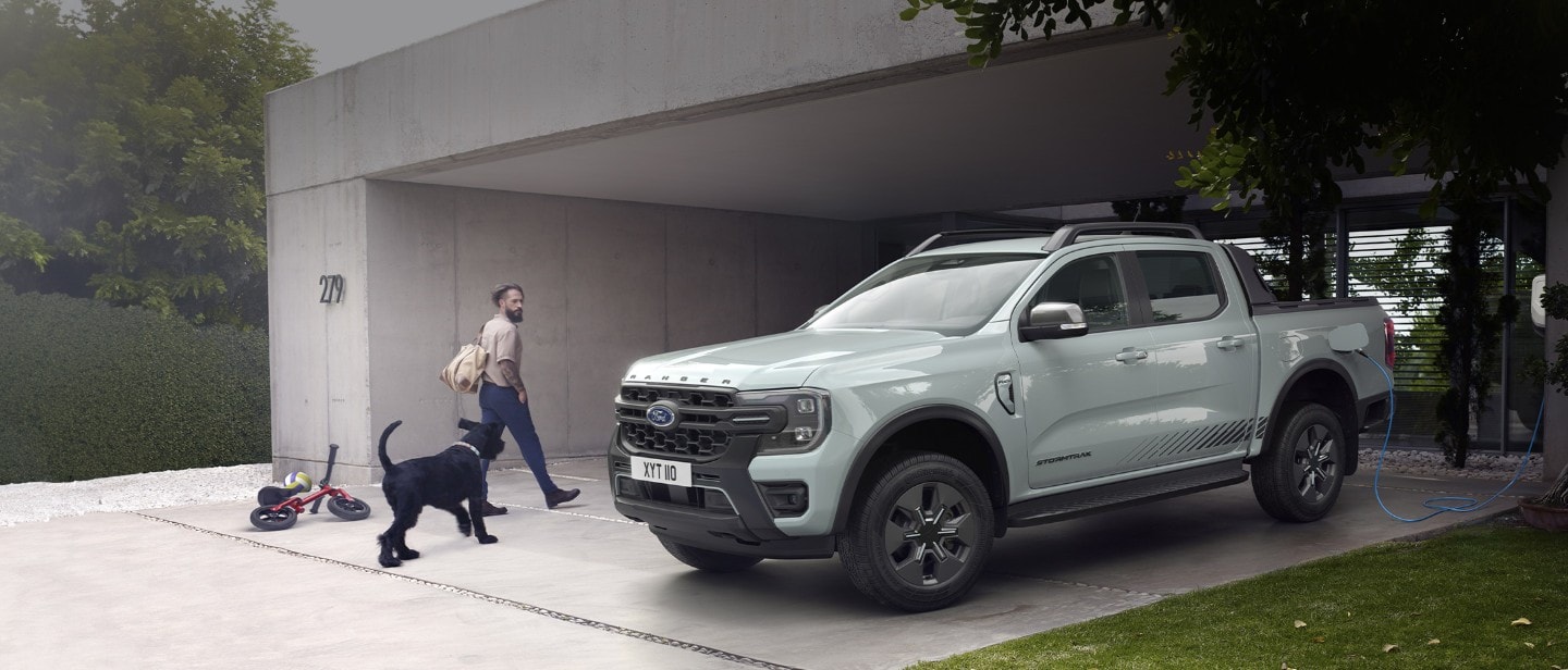 Een man en zijn hond gaan een modern huis binnen en wandelen langs een geparkeerde Ford Ranger Stormtrak Plug-in Hybrid die wordt opgeladen. 