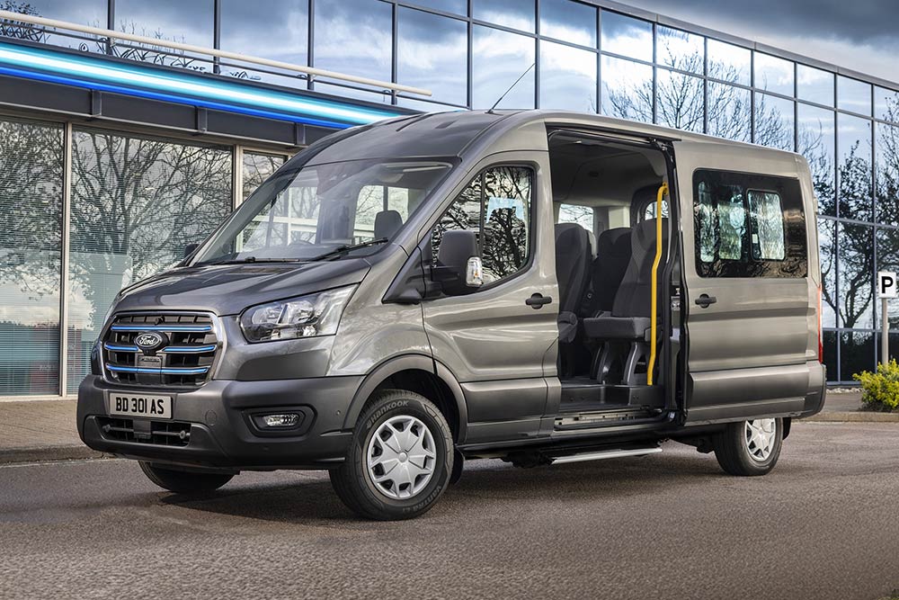 Zijaanzicht van een grijze Ford Pro Transit met open deuren, geparkeerd voor een modern gebouw.