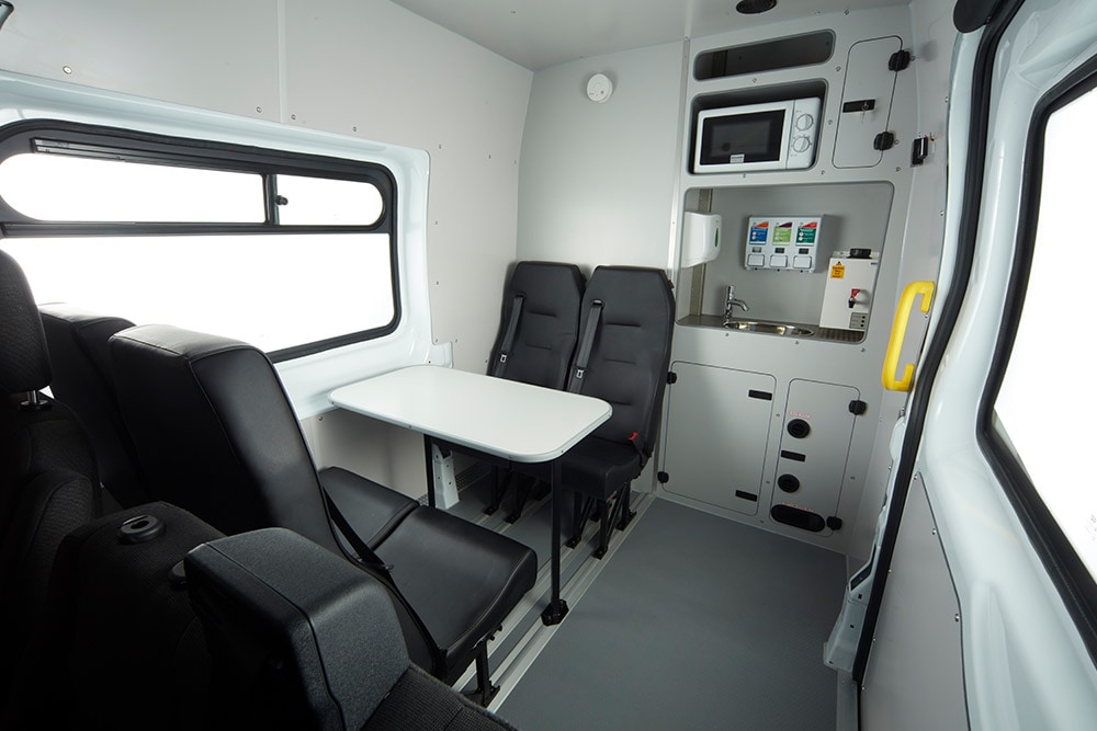 Interieur van een Ford bestelwagen, met een tafel, zwarte stoelen en een compacte kitchenette met een magnetron.