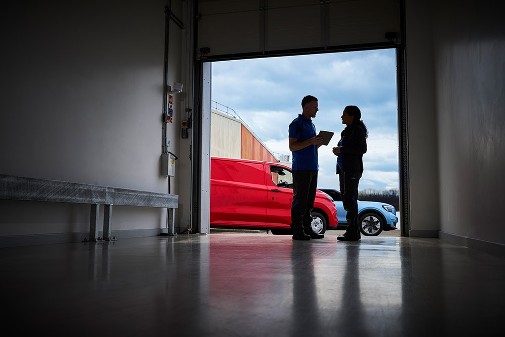 Twee mensen praten in een garagedeur, met een rode Ford-bestelwagen en een blauwe Ford SUV buiten geparkeerd.