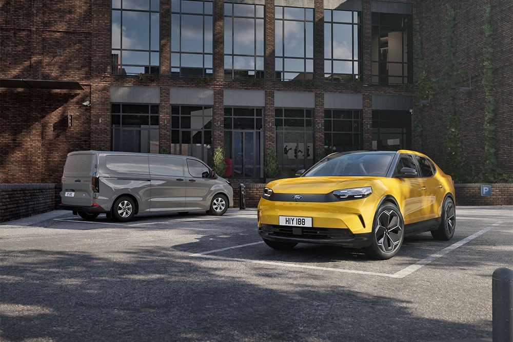  Een grijze Ford-bestelwagen en een gele Capri geparkeerd voor een modern bakstenen gebouw.