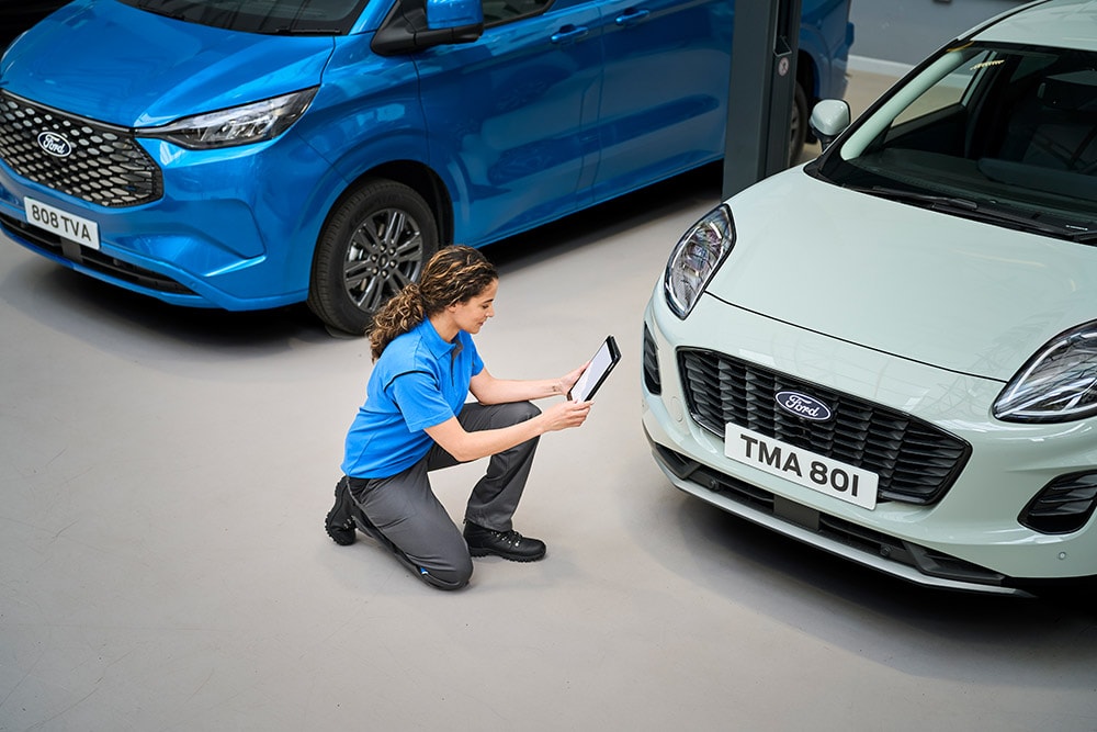 Een technicus knielt tussen een blauwe Ford-bestelwagen en een witte Ford Puma en kijkt naar een tablet.