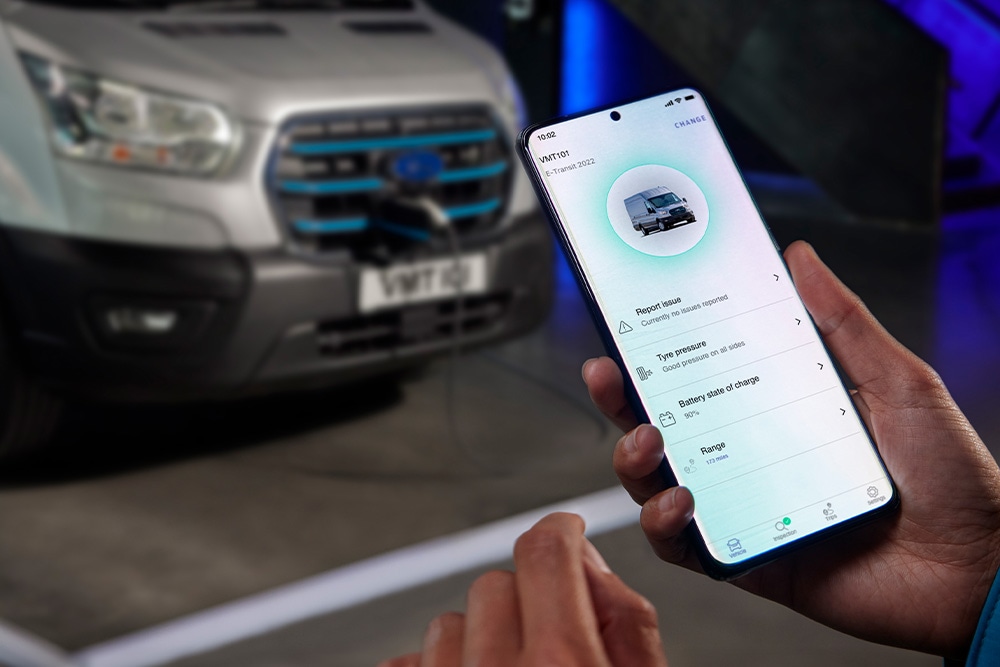 Een persoon houdt een smartphone vast met een Ford-app, terwijl een Ford E-Transit Custom op de achtergrond oplaadt.