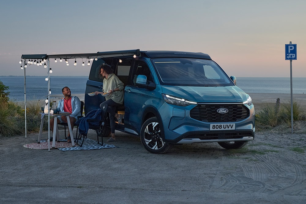 Twee kampeerders ontspannen bij een blauwe Ford Pro Transit Custom Nugget, een parkeerplaats aan het strand op de achtergrond.
