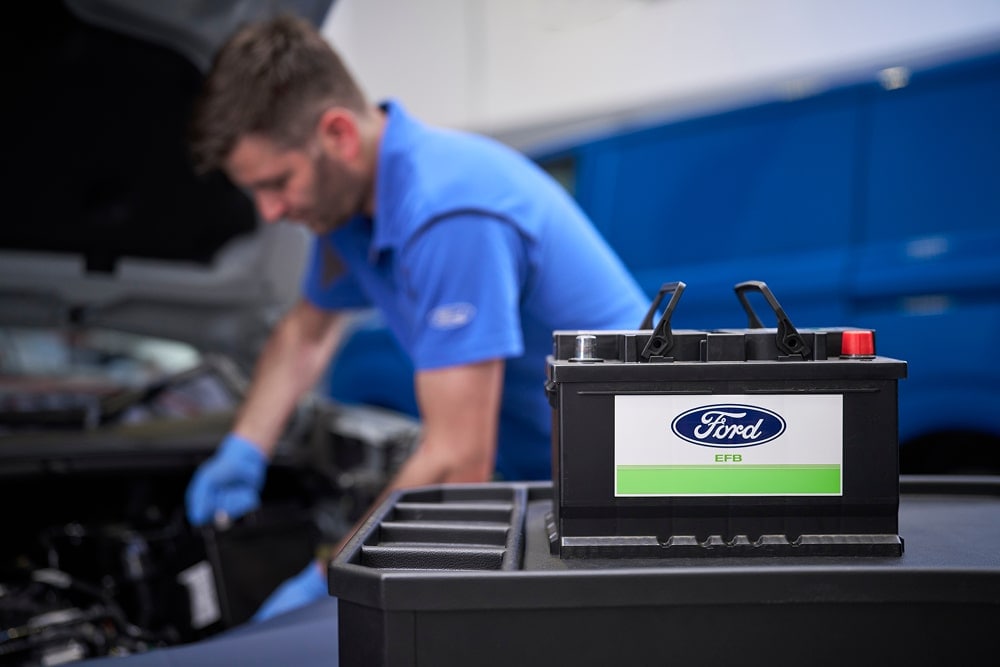 Een Ford-monteur werkt aan een automotor, met een Ford-autobatterij op de voorgrond.