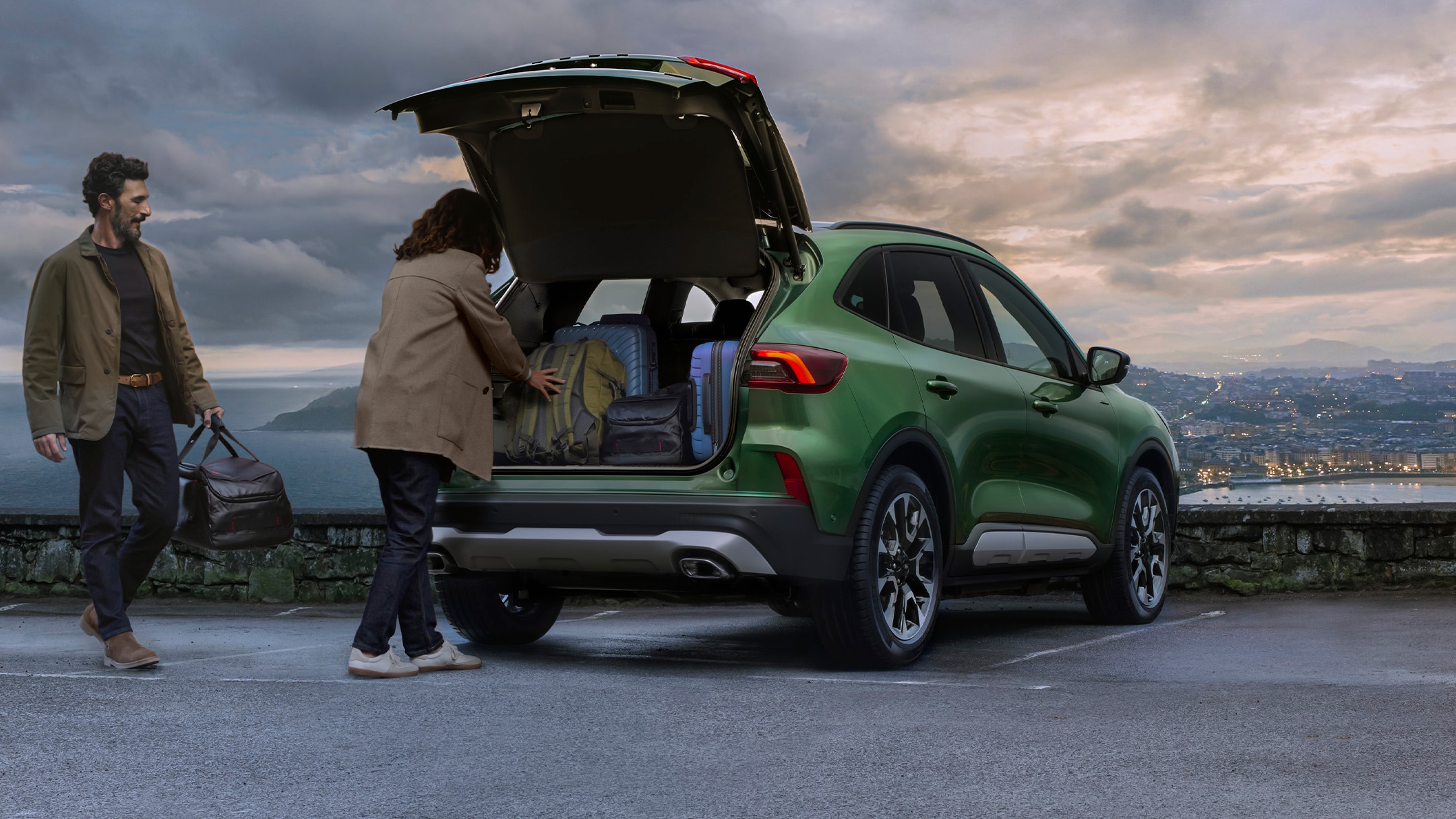 Een man en een vrouw laden bagage in de kofferbak van een groene SUV die geparkeerd staat op een verhard uitkijkpunt.